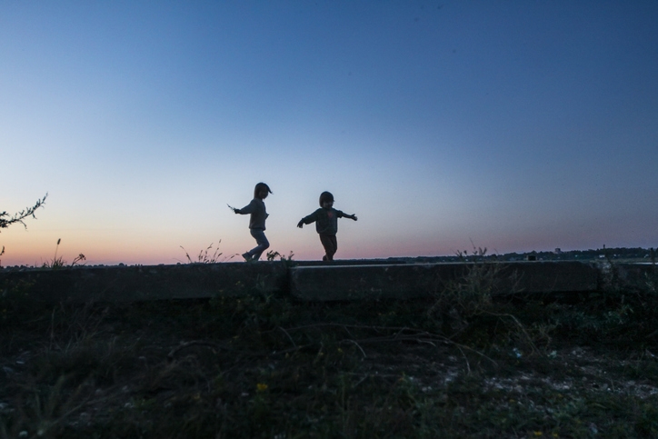 Two children silhouetted against the evening sky, joyfully running on a grassy hill
