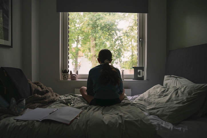 Person sitting on a bed, looking out a window at trees. The scene conveys contemplation or relaxation. Bedroom has an open notebook nearby