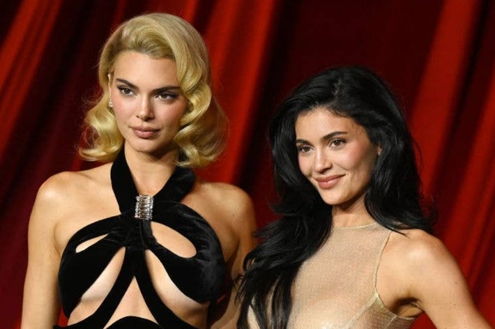 Two women on a red carpet, one in a black cut-out gown with a silver accent and wavy hair, the other in a beige sheer dress with long dark hair