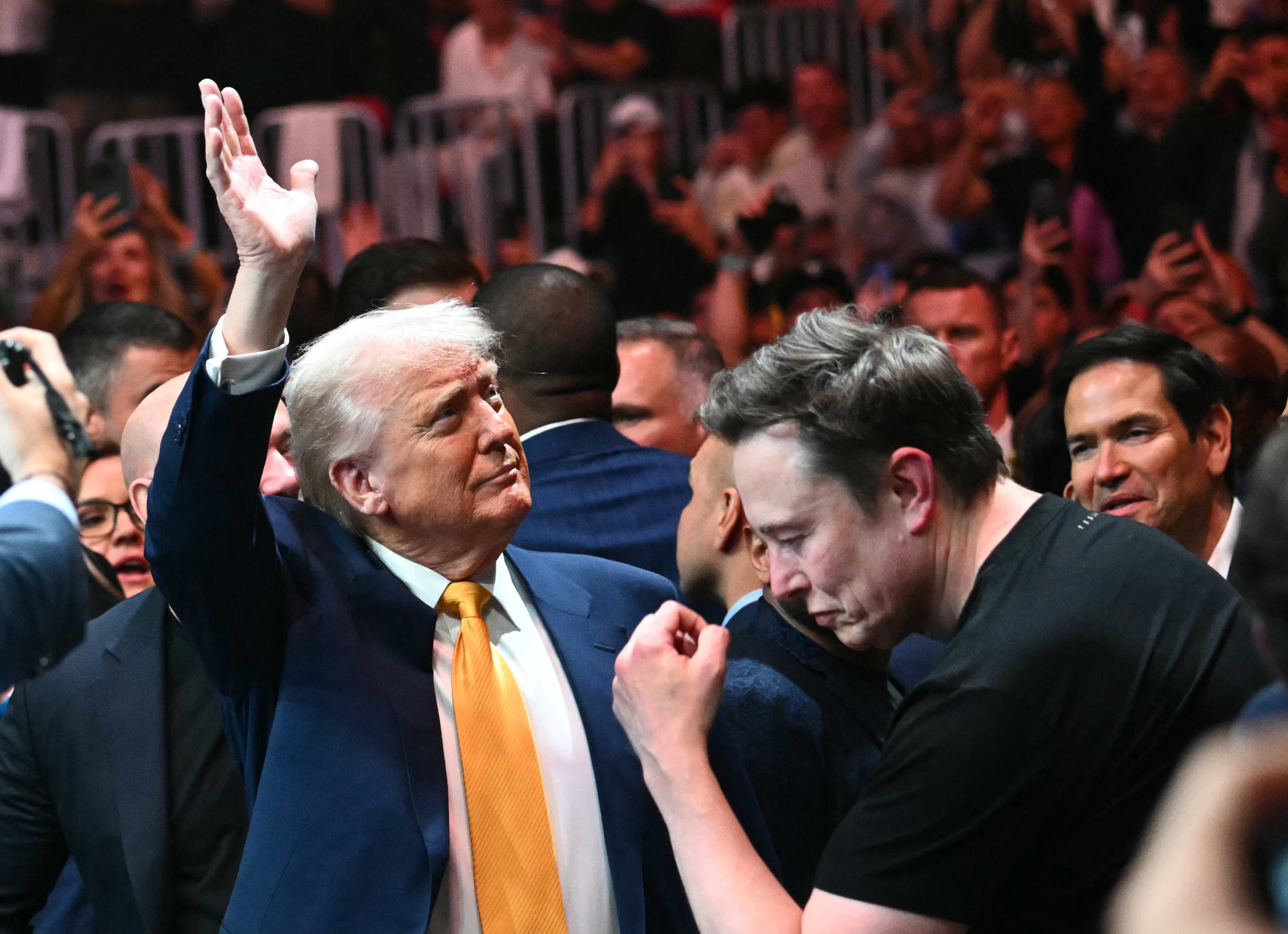Two people greet each other at a crowded event, one in a suit and the other in a casual T-shirt