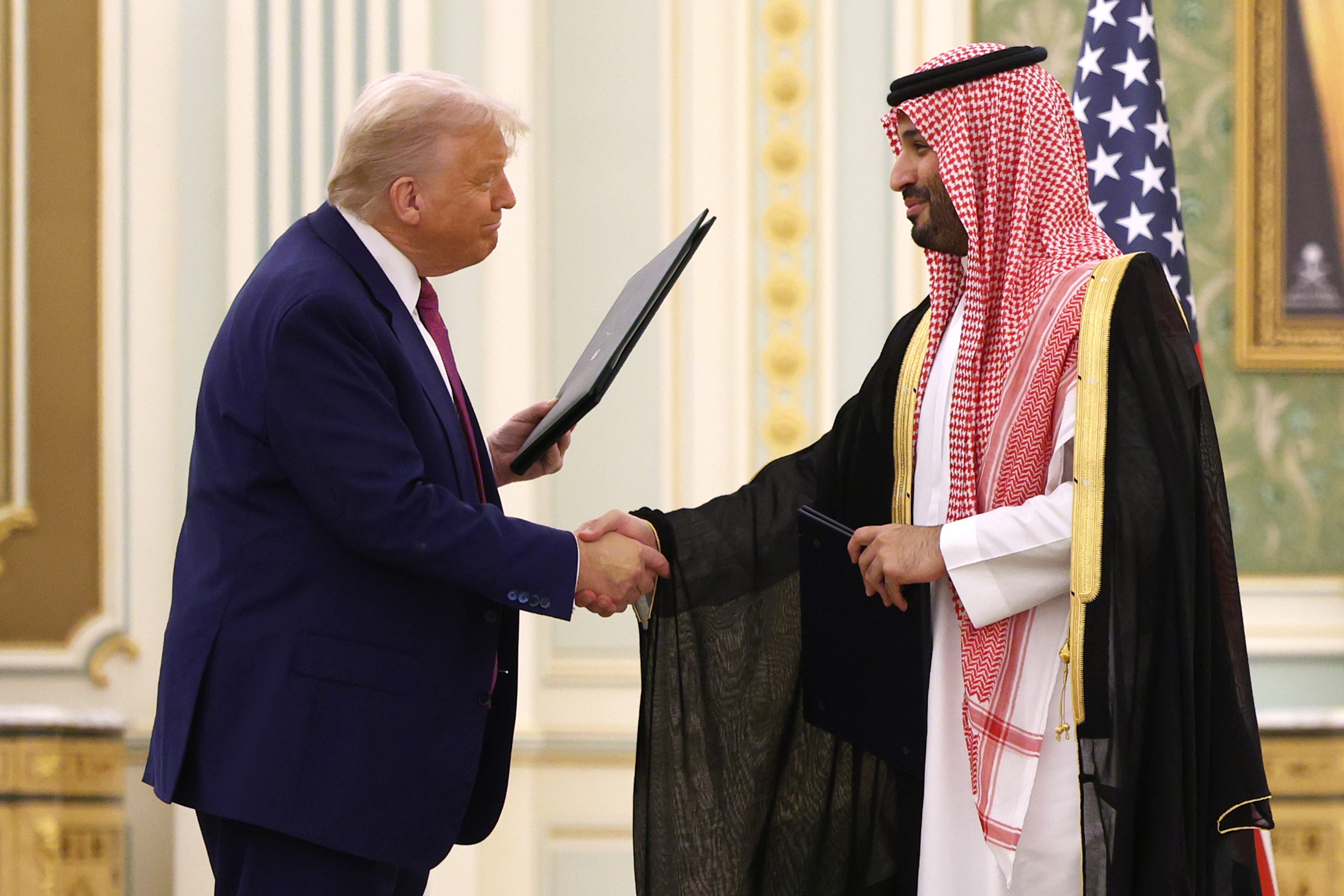 Two men shaking hands; one in a suit, the other in traditional attire, holding a document in an ornate room
