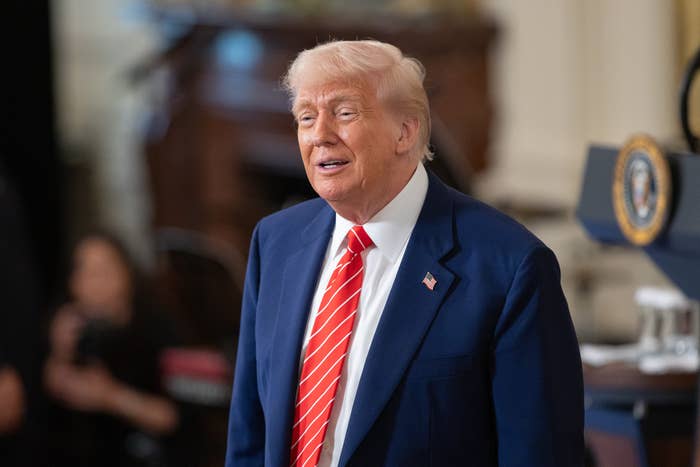 A person in a suit and striped tie stands in front of a podium with a presidential seal, smiling