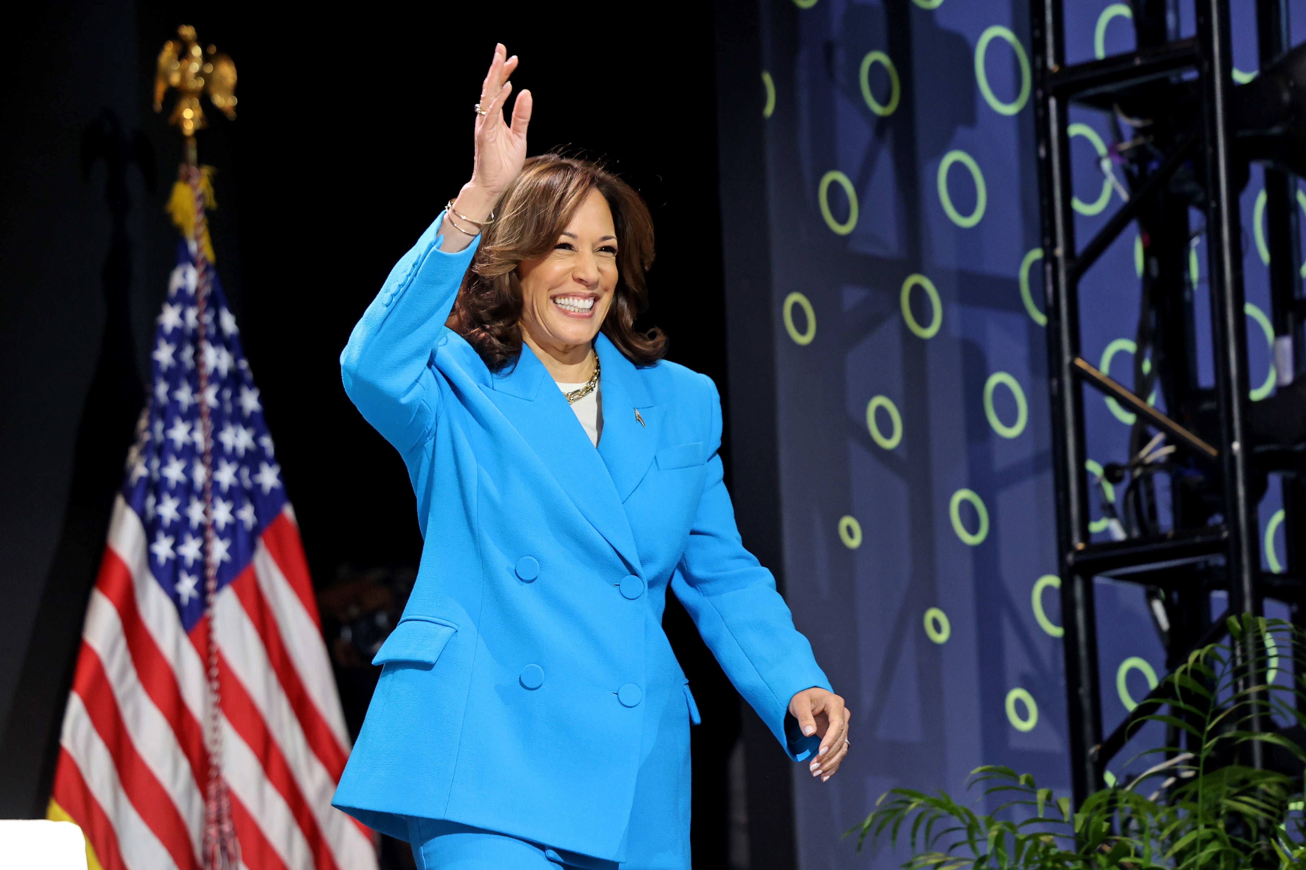 Kamala Harris waves while walking onstage, wearing a well-tailored pantsuit, with a U.S. flag and patterned backdrop behind them