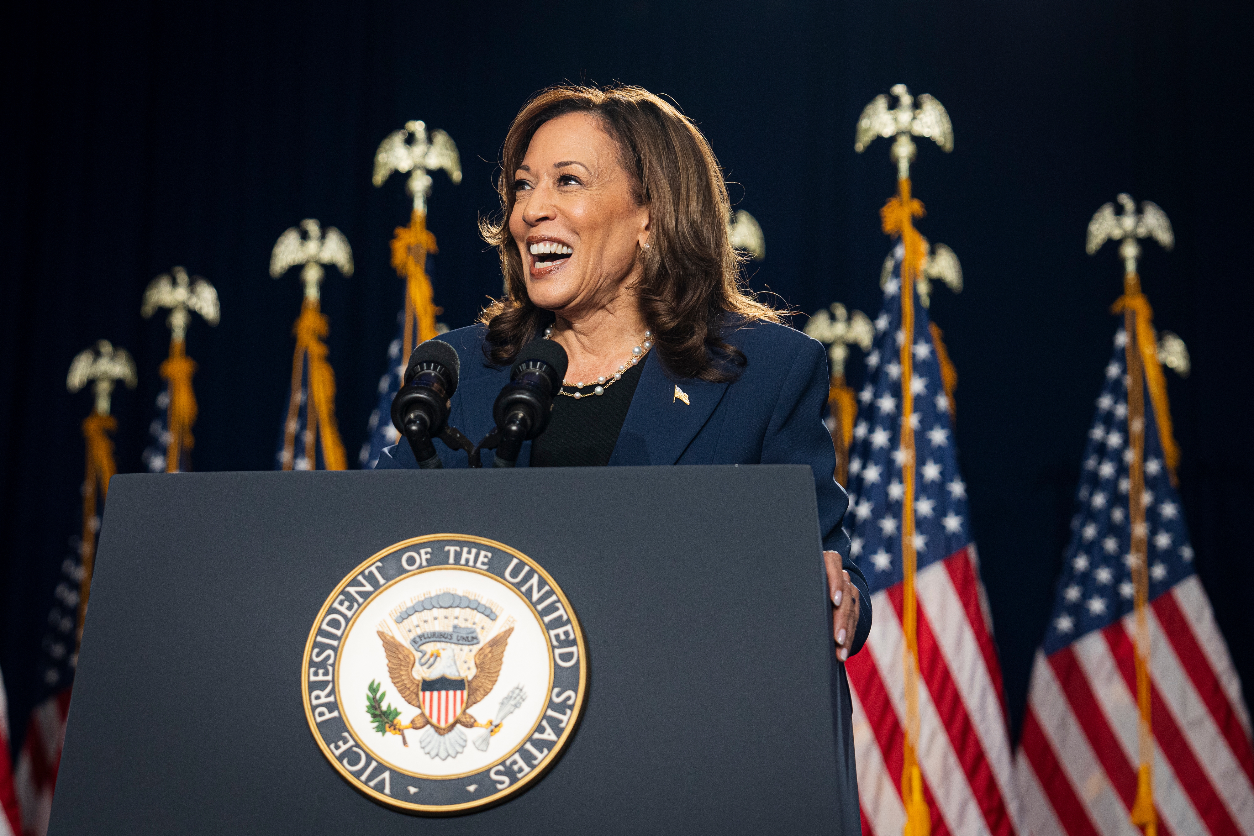 Kamala Harris stands smiling at a podium with microphones, the U.S. Vice Presidential seal, and several American flags in the background
