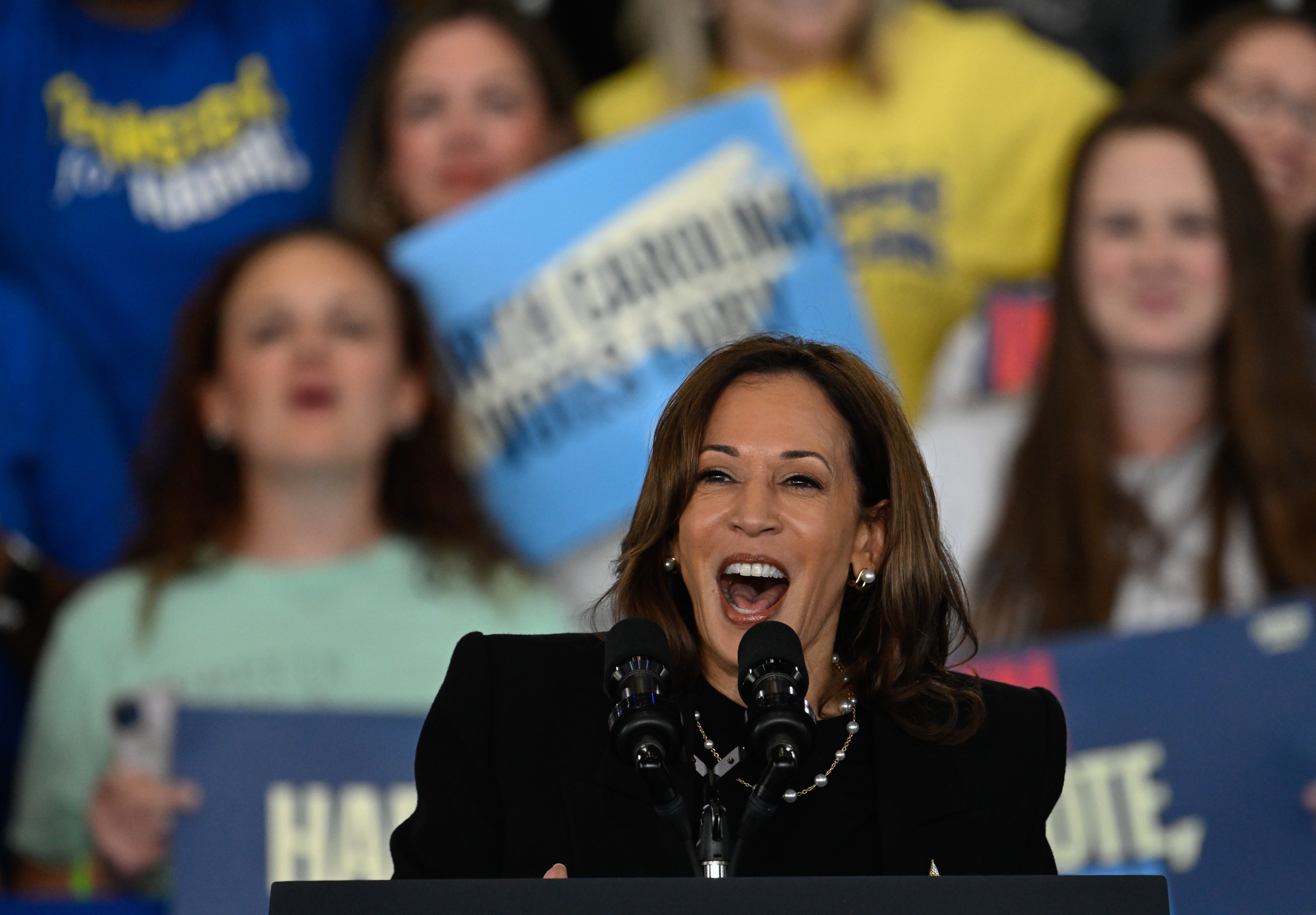 Kamala Harris speaks energetically at a podium with microphones, surrounded by a crowd holding signs, creating an engaging atmosphere