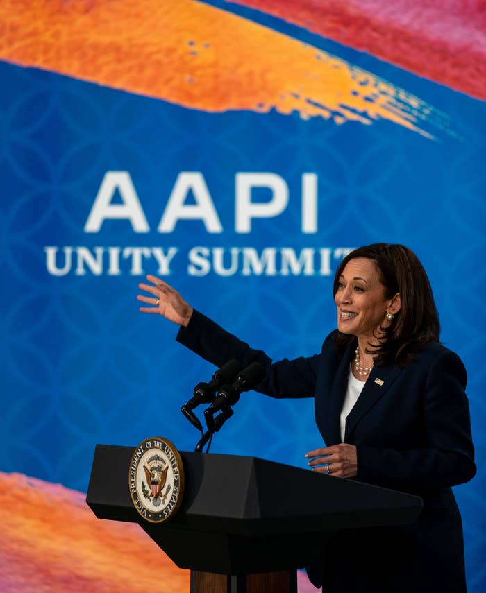Kamala Harris gestures while addressing the AAPI Unity Summit from a podium with the presidential seal. A vibrant abstract background is behind them