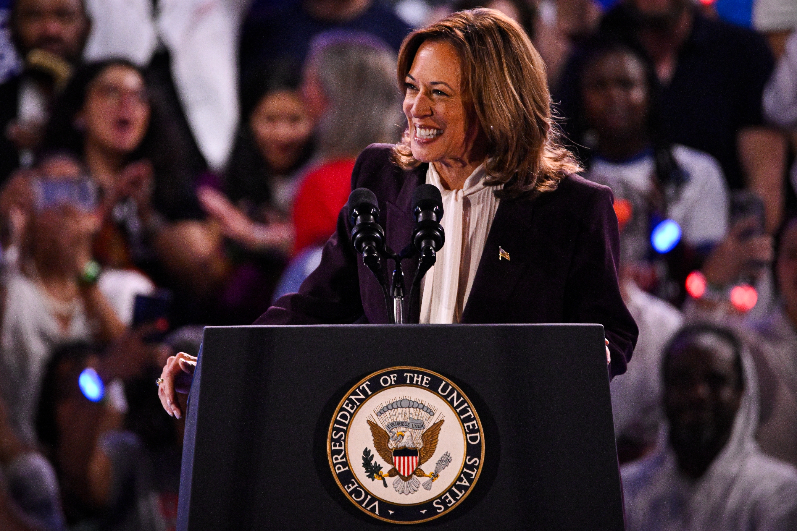 A person stands behind a podium with the presidential seal, smiling, while speaking to a crowd