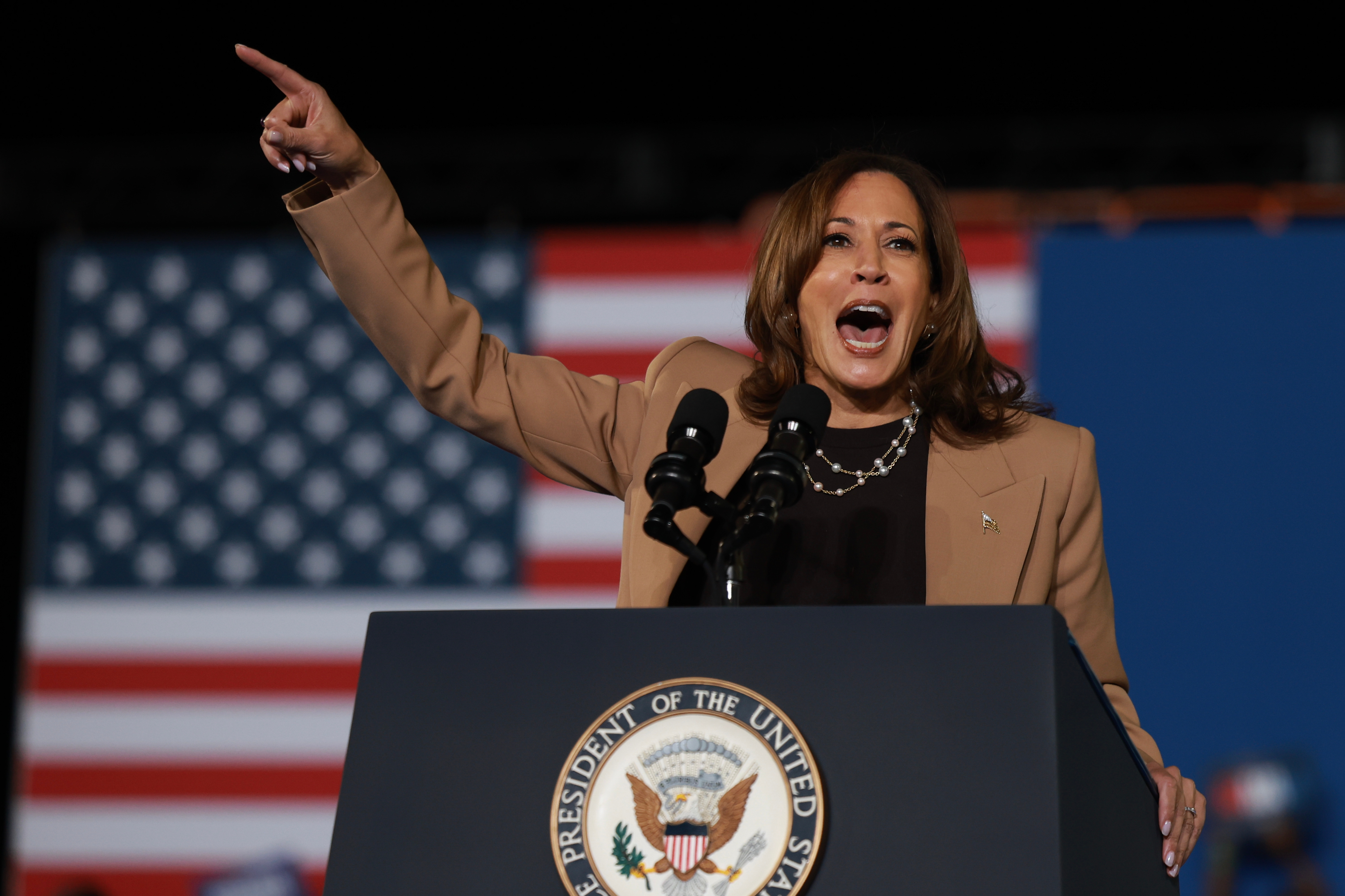 Person speaking passionately at a podium with microphones, hand raised, in front of a blurred American flag backdrop
