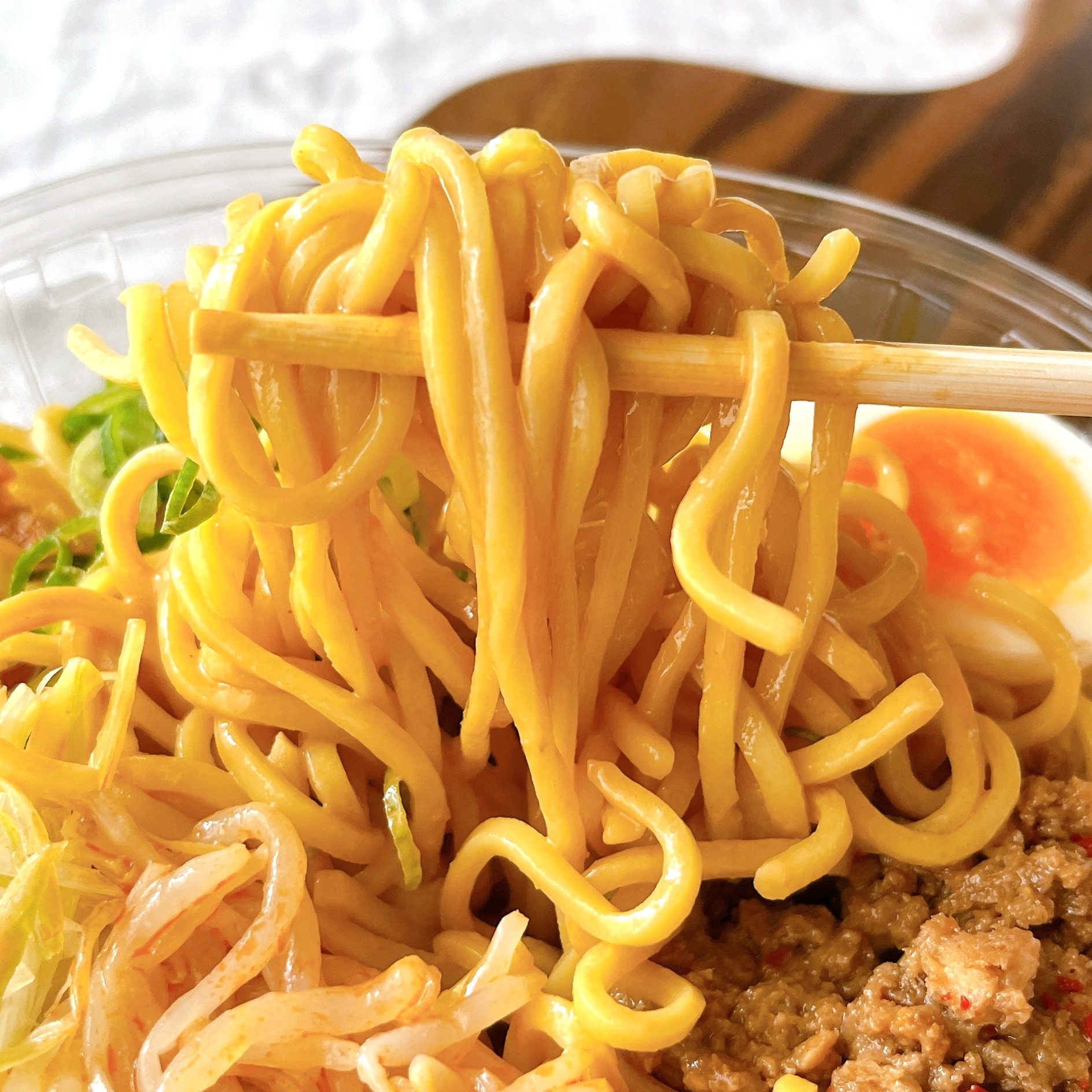 麺を箸で持ち上げたラーメンのクローズアップ。野菜や肉、半熟卵がトッピングされています。