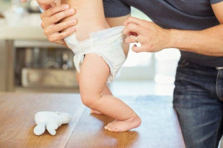 男性がキッチンで赤ちゃんのオムツをのぞきながらミルクを飲ませている。親しみやすい家族のひととき。