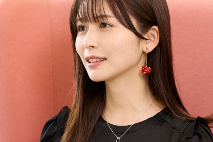 女性が椅子に座って微笑んでいる。黒いトップスに赤いイヤリングを着用し、プロフェッショナルな雰囲気。