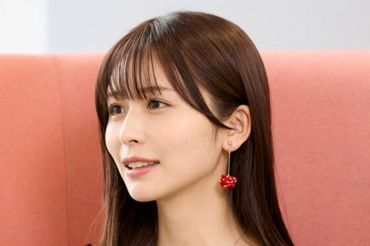 女性が椅子に座って微笑んでいる。黒いトップスに赤いイヤリングを着用し、プロフェッショナルな雰囲気。