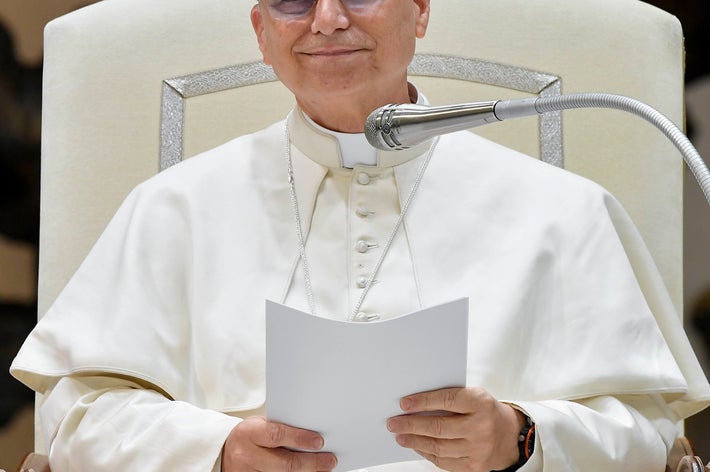 白い服装の男性が椅子に座り、マイクの前で紙を持っている。