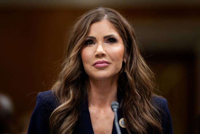 A person with long wavy hair, in formal business attire, speaks at a public hearing