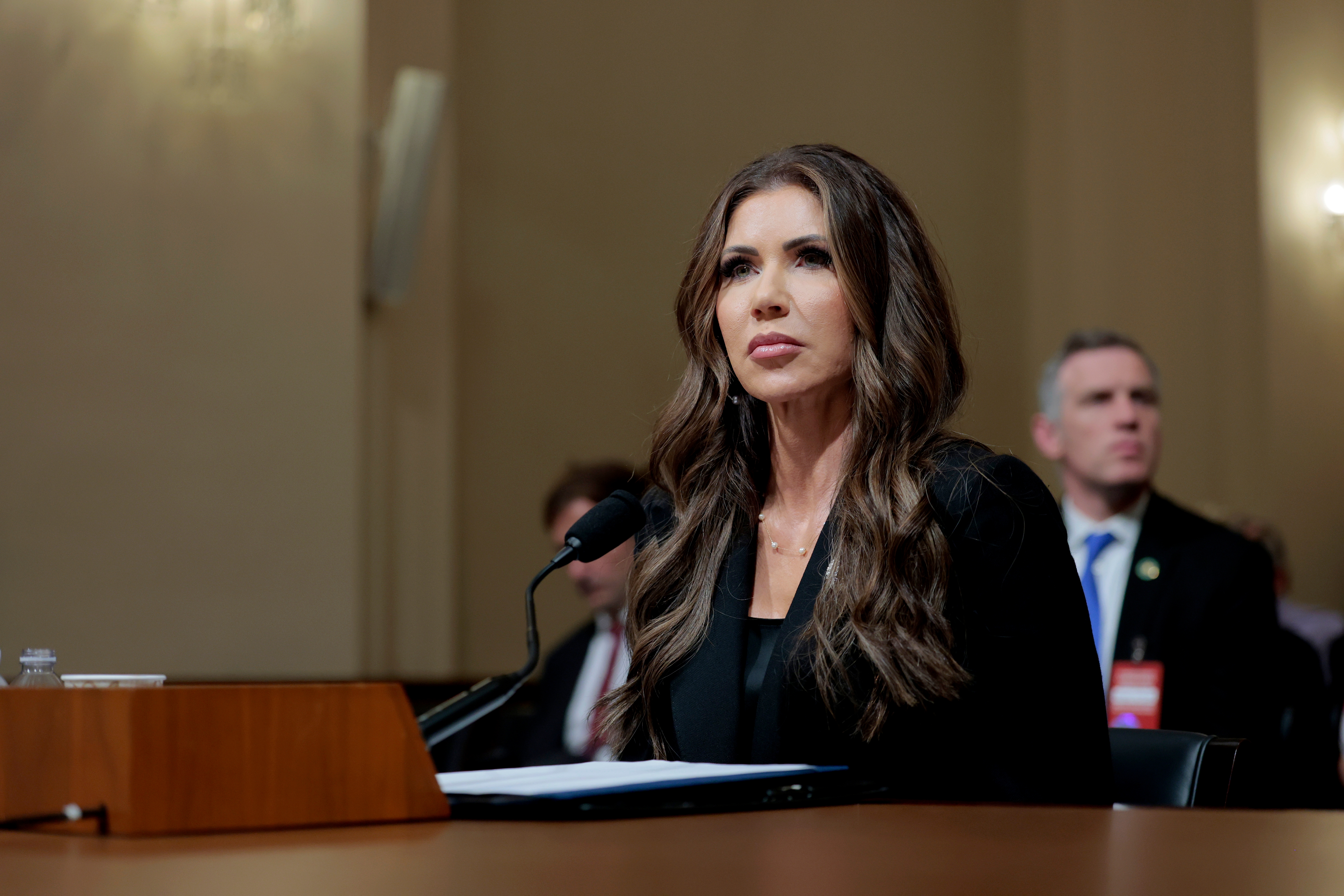 Woman with long hair testifying at a hearing, sitting at a desk with a microphone and paper, focused expression, audience members in the background