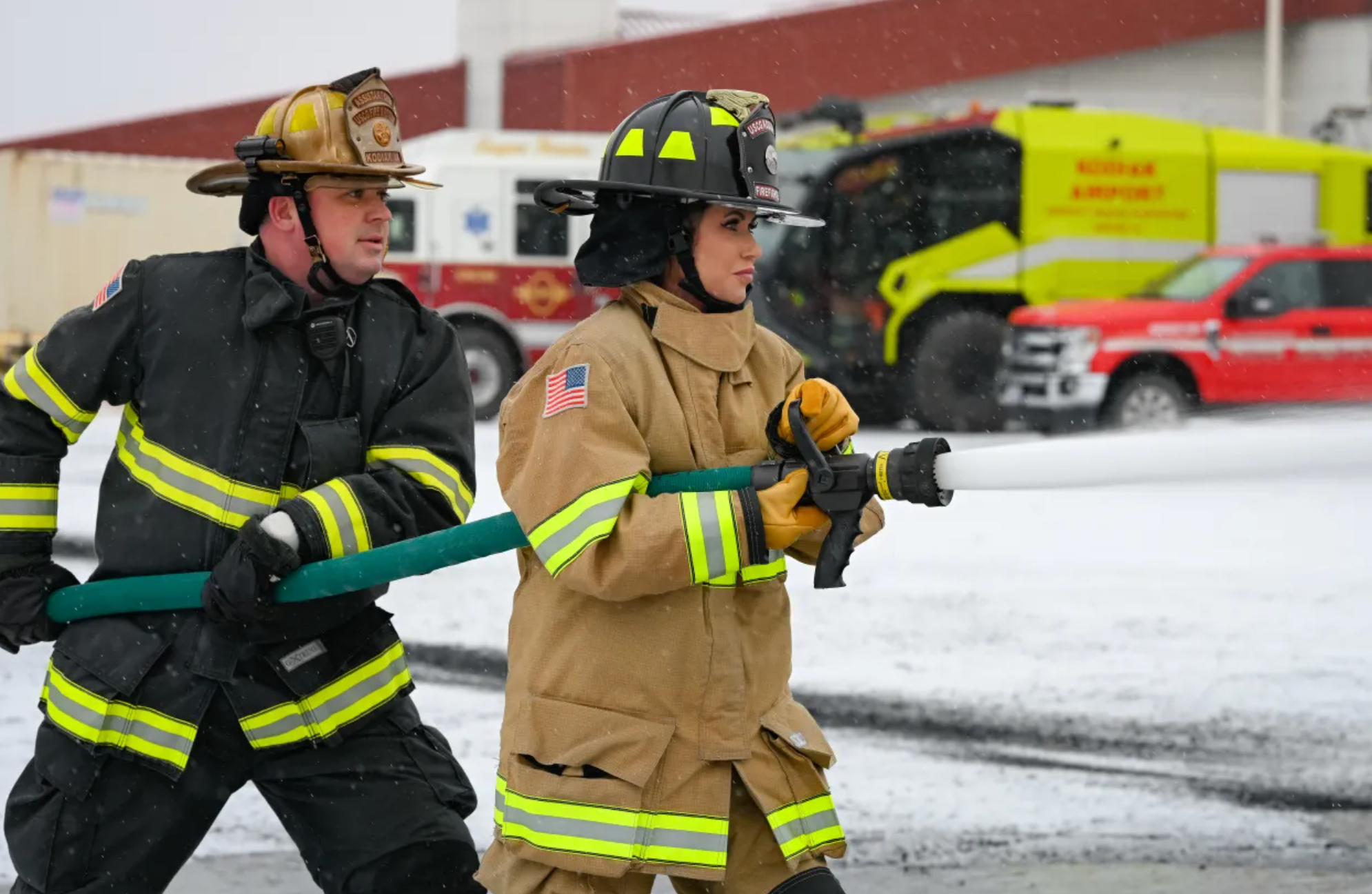 Firefighters in full gear are actively handling a hose during a snowy emergency response, with fire trucks visible in the background