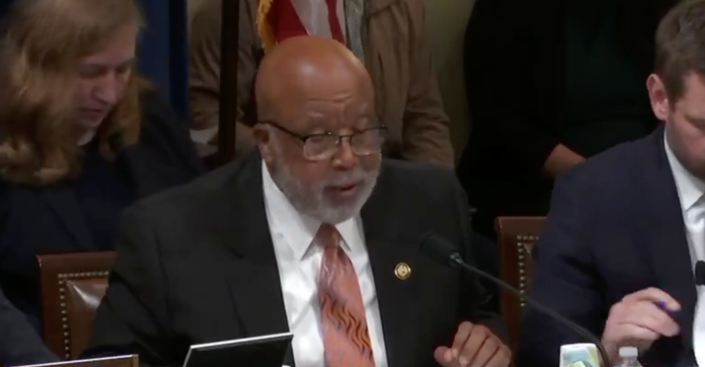 A person in a suit speaks at a meeting, seated at a table with microphones and papers, with others present