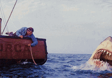 A man on a boat marked "Orca" reaches toward a large shark emerging from the water, capturing a dramatic moment from a famous movie scene