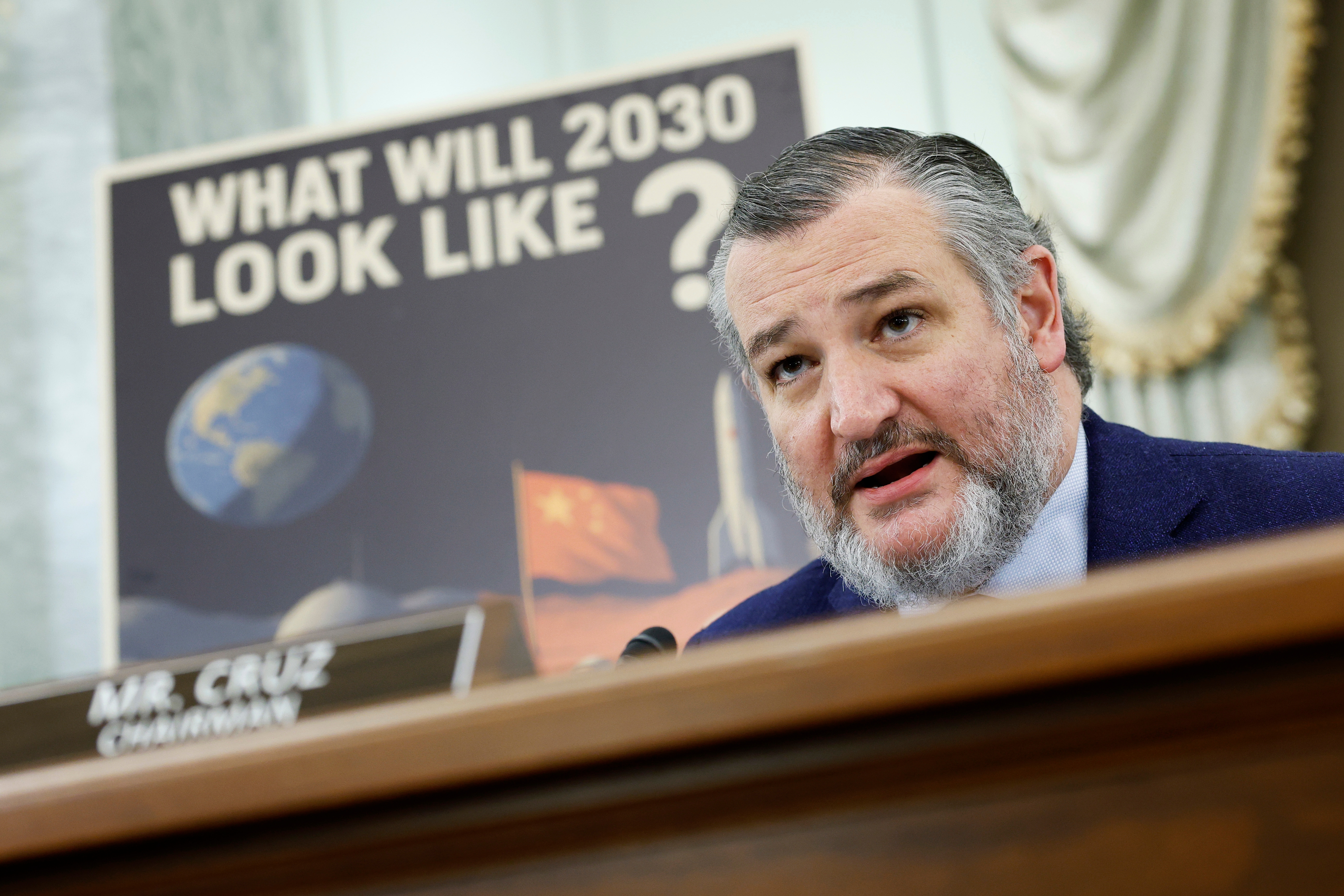 A person speaks at a podium labeled "MR. CRUZ CHAIRMAN" in front of a poster questioning the future in 2030, featuring global imagery