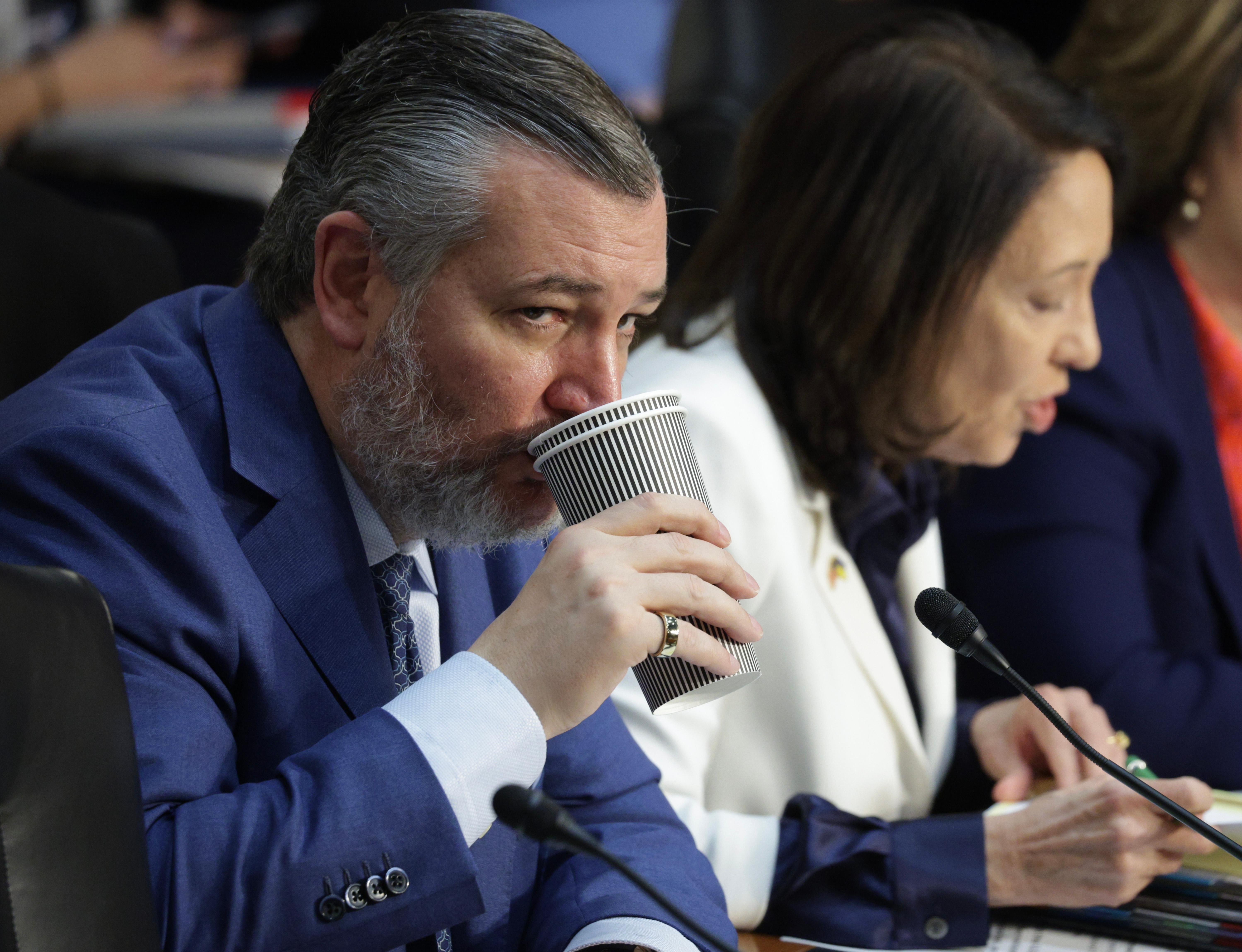 Two people sit at a table with microphones. The man, in a suit, drinks from a striped cup. The woman, in a white jacket, appears to be speaking