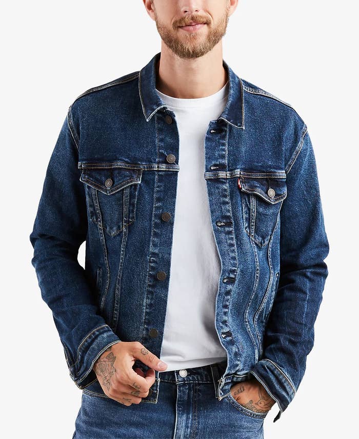 Pria mengenakan jaket denim di atas kaus putih, dengan tato di tangannya, menampilkan gaya kasual untuk artikel yang berhubungan dengan perbelanjaan