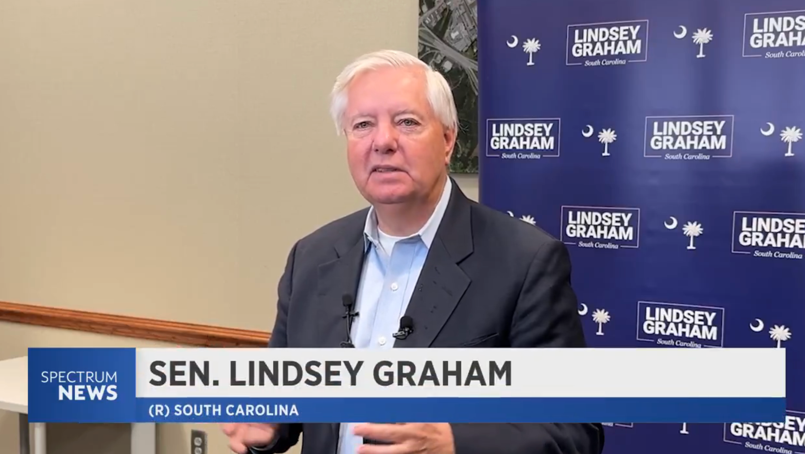 A man in a suit stands before a backdrop covered with "Lindsey Graham" text; he seems to be speaking on camera
