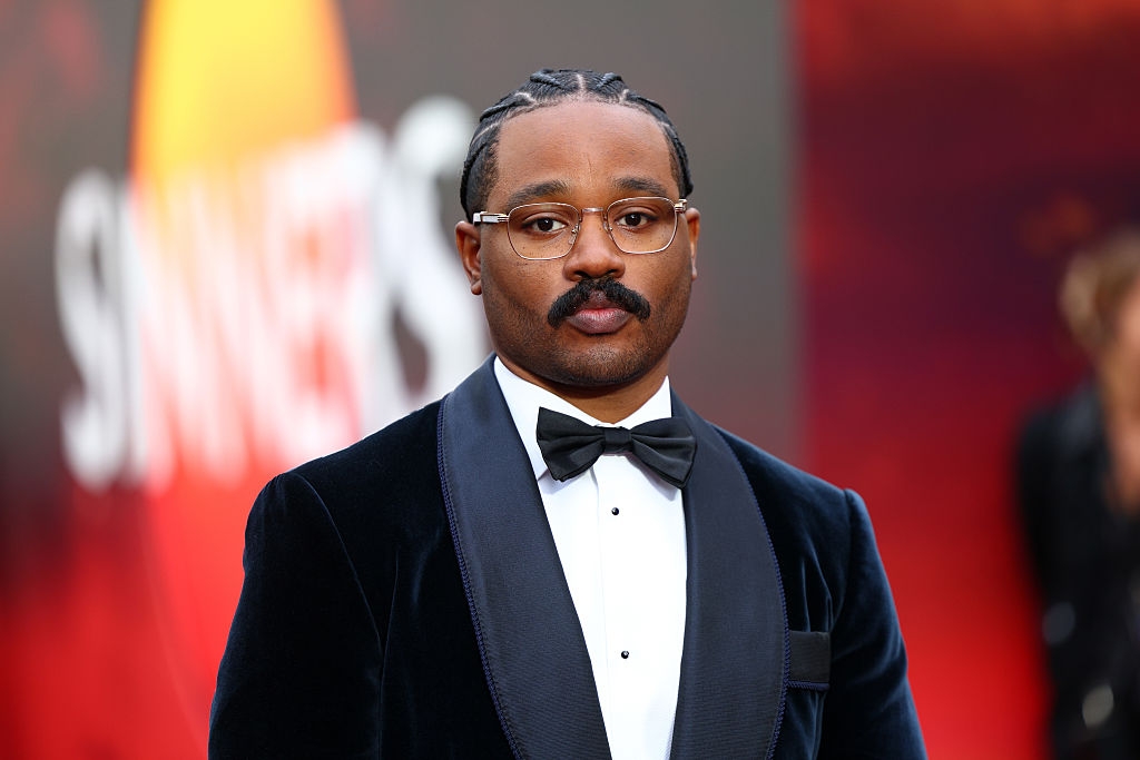 A person on a red carpet wearing a velvet tuxedo with a bow tie and glasses, standing in front of an event backdrop
