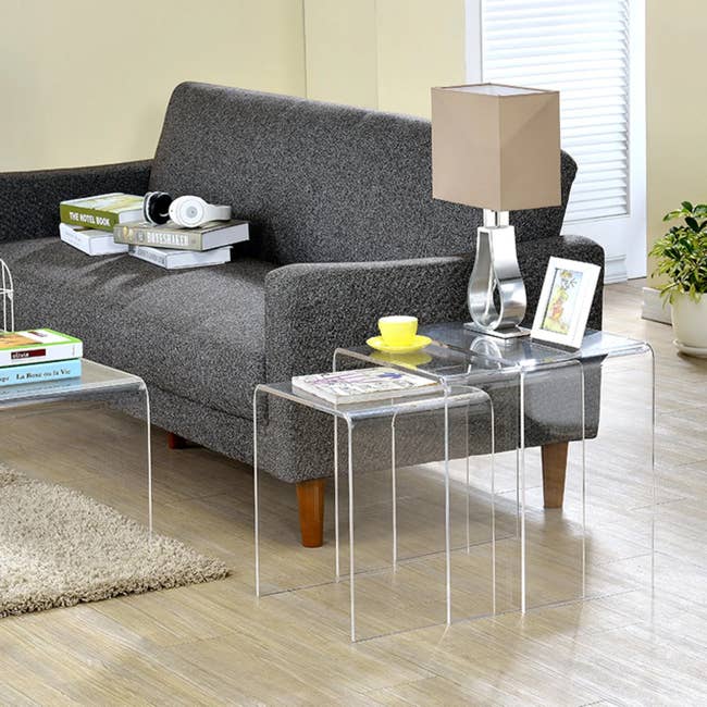 Living country with a grey sofa, wide nesting tables, a lamp, a stack of books, a teacup connected a saucer, and a framed photo