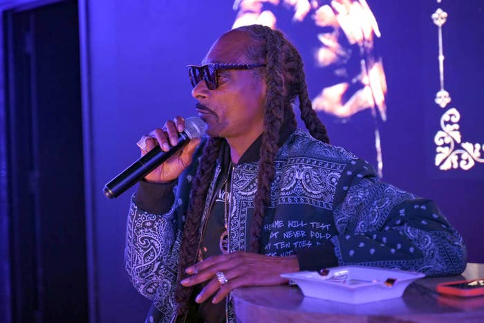Snoop Dogg with long braids and sunglasses speaks into a microphone, wearing a patterned jacket, seated at a table with a notebook