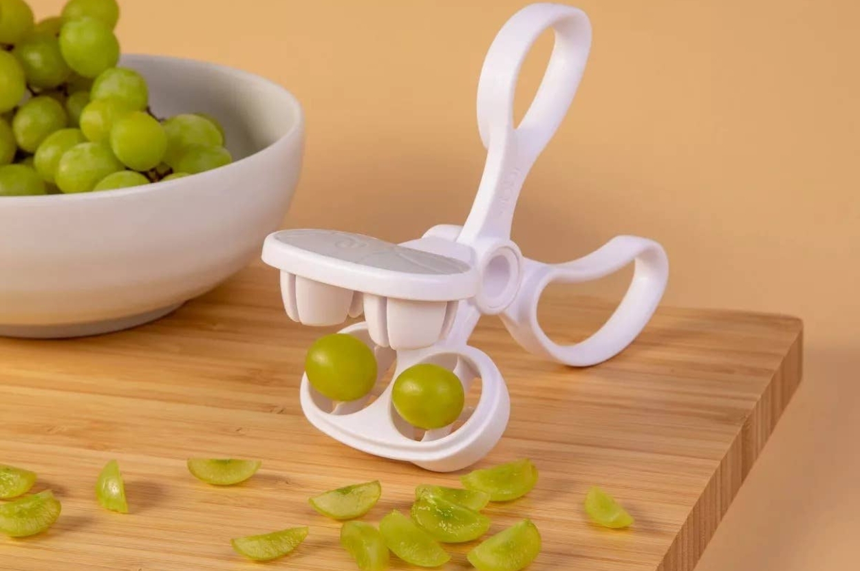 Grape slicer with two grapes ready to slice, surrounded by cut grape pieces, on a wooden board near a bowl of whole grapes