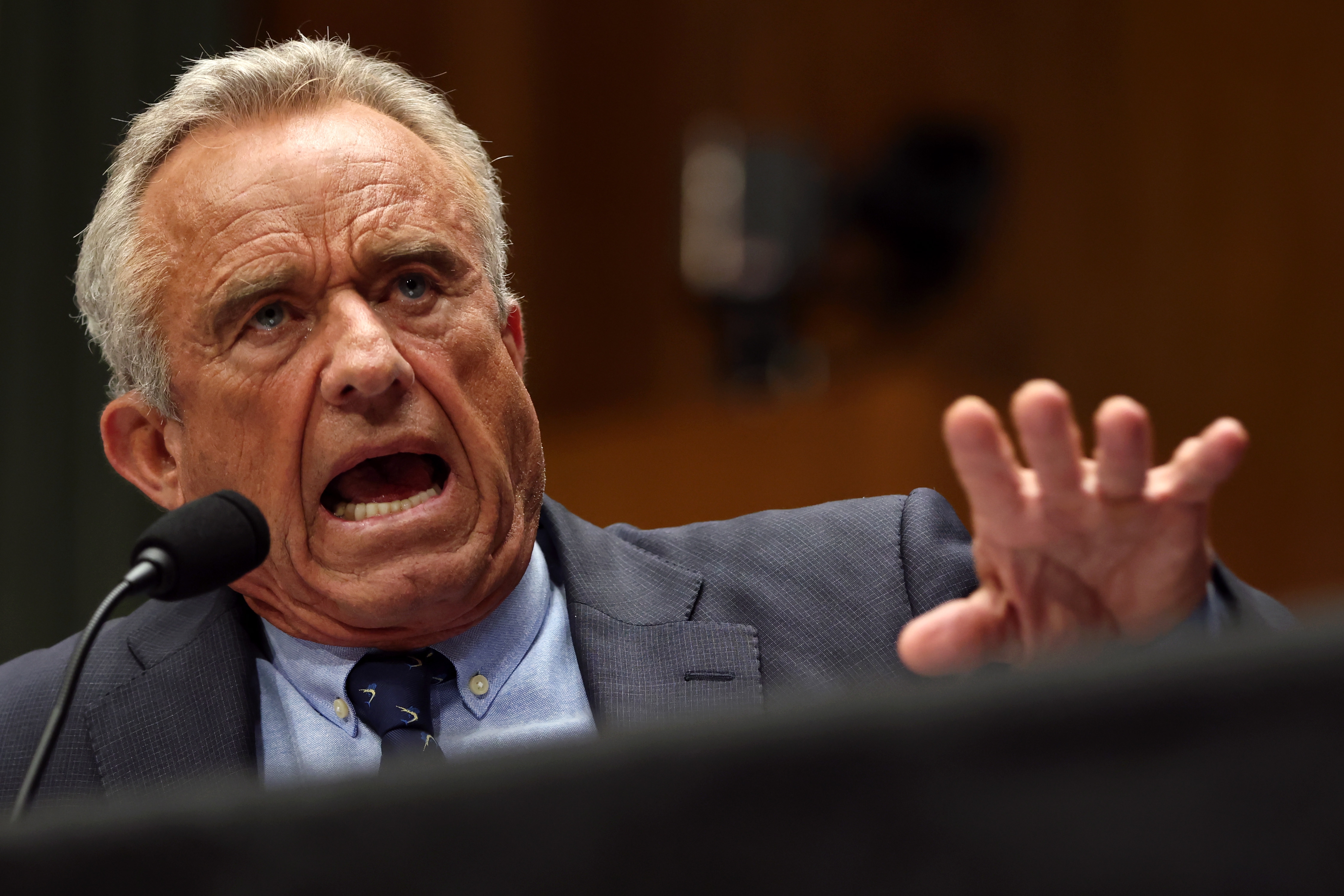 A person speaks passionately at a podium, wearing a suit and tie, gesturing with one hand