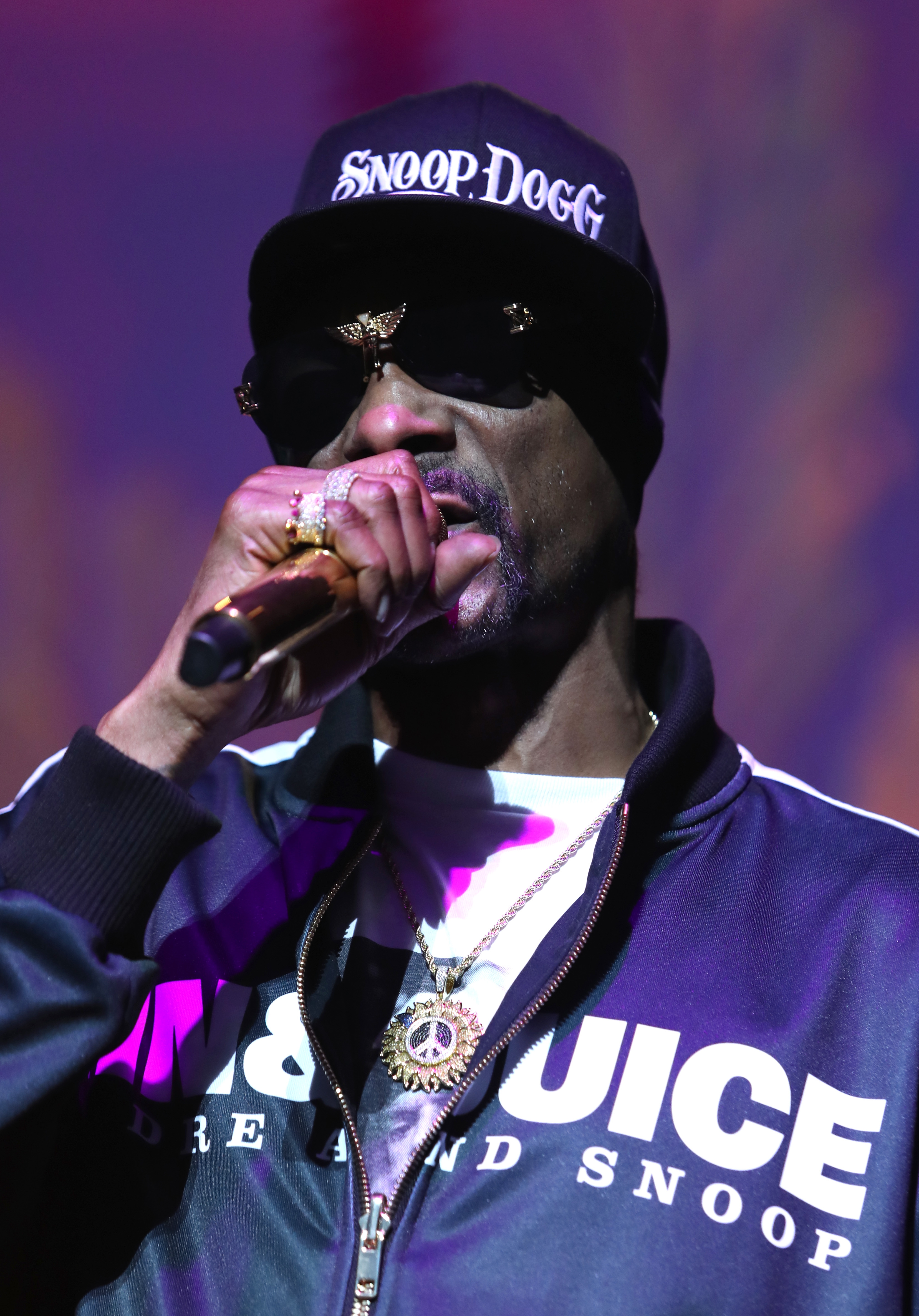 Snoop Dogg in a track jacket, sunglasses, and cap performs onstage, holding a microphone
