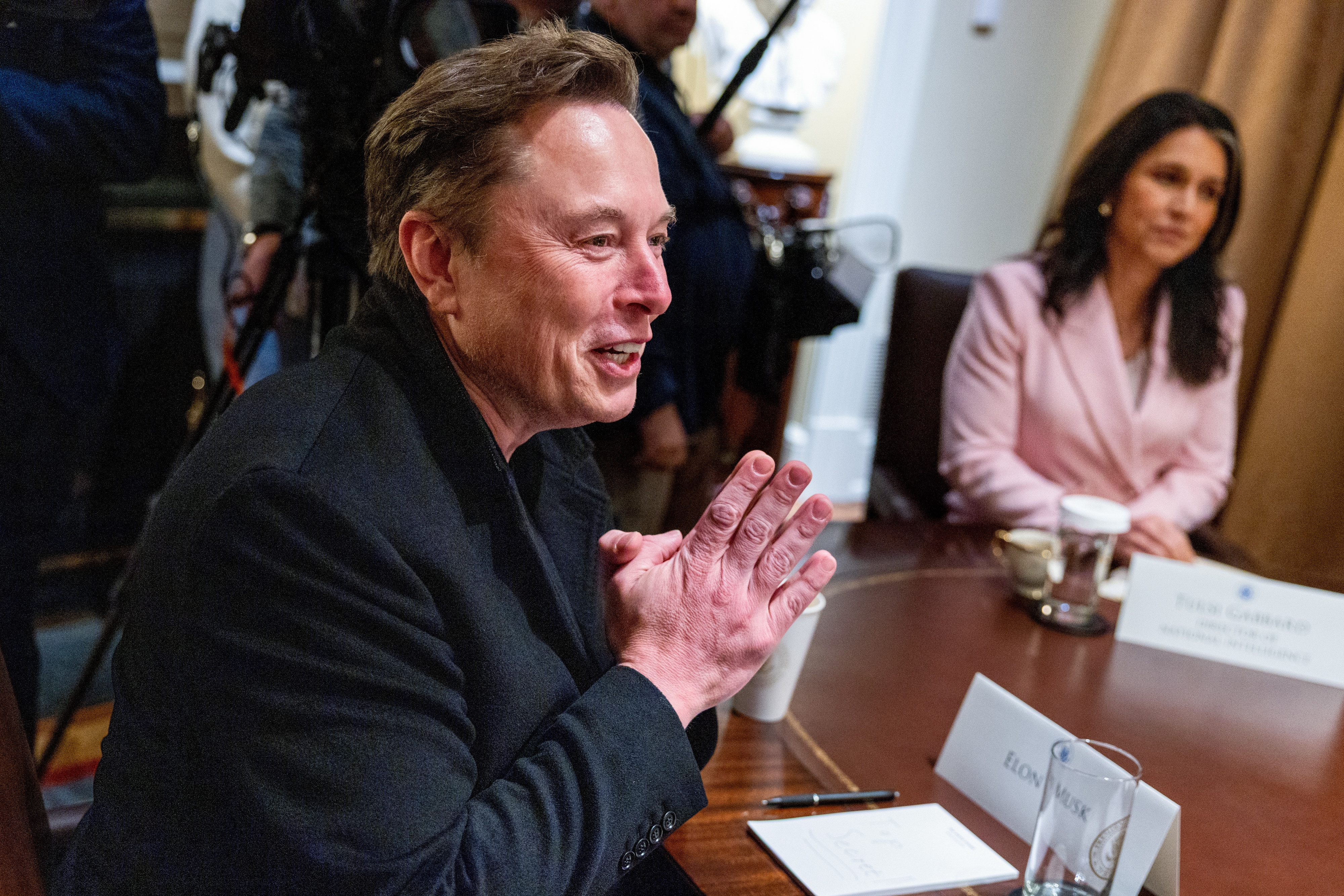 A person in a suit is seated at a meeting table, speaking animatedly with another person in professional attire in the background
