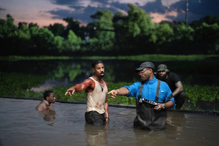 Actor in a muddy tank, discussing a scene with crew members while filming. Surrounded by water and foliage