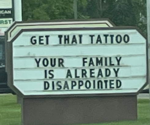 Sign reads: "Get that tattoo. Your family is already disappointed." Nearby, a pedestrian signal shows a stop hand