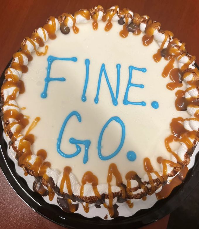 A cake with decorative caramel drizzle and chocolate chips features icing text saying "FINE. GO."