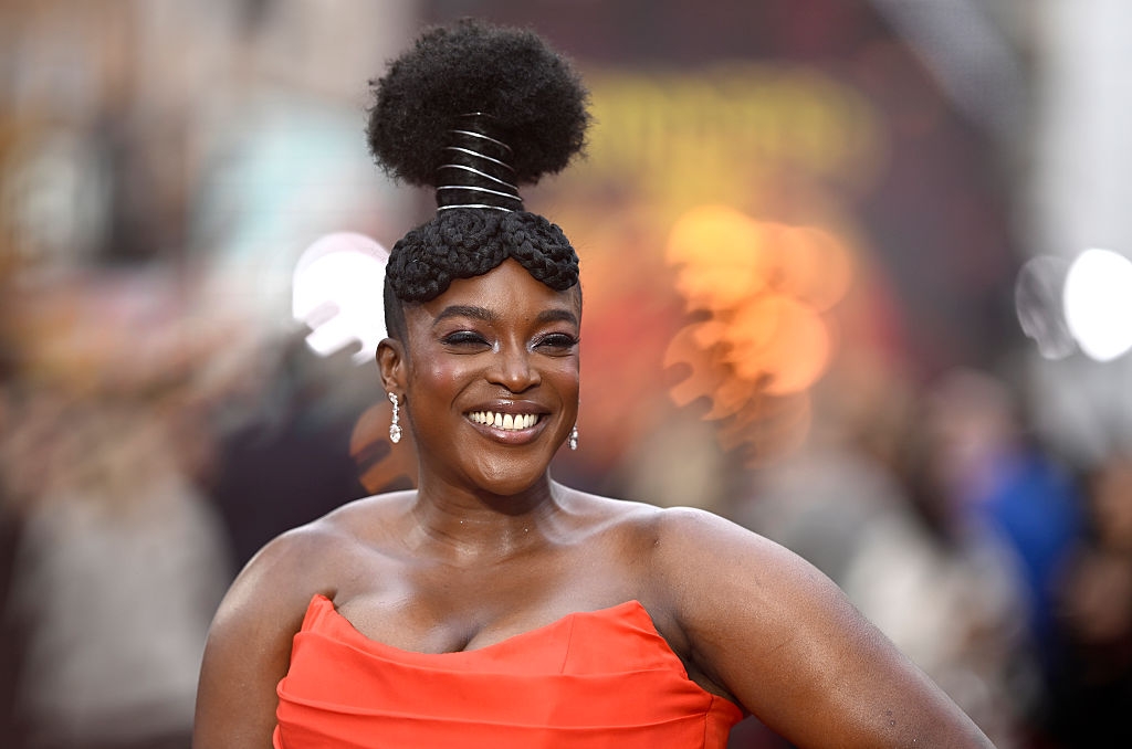Person smiling on red carpet, wearing a strapless gown and unique hairstyle with an elaborate design