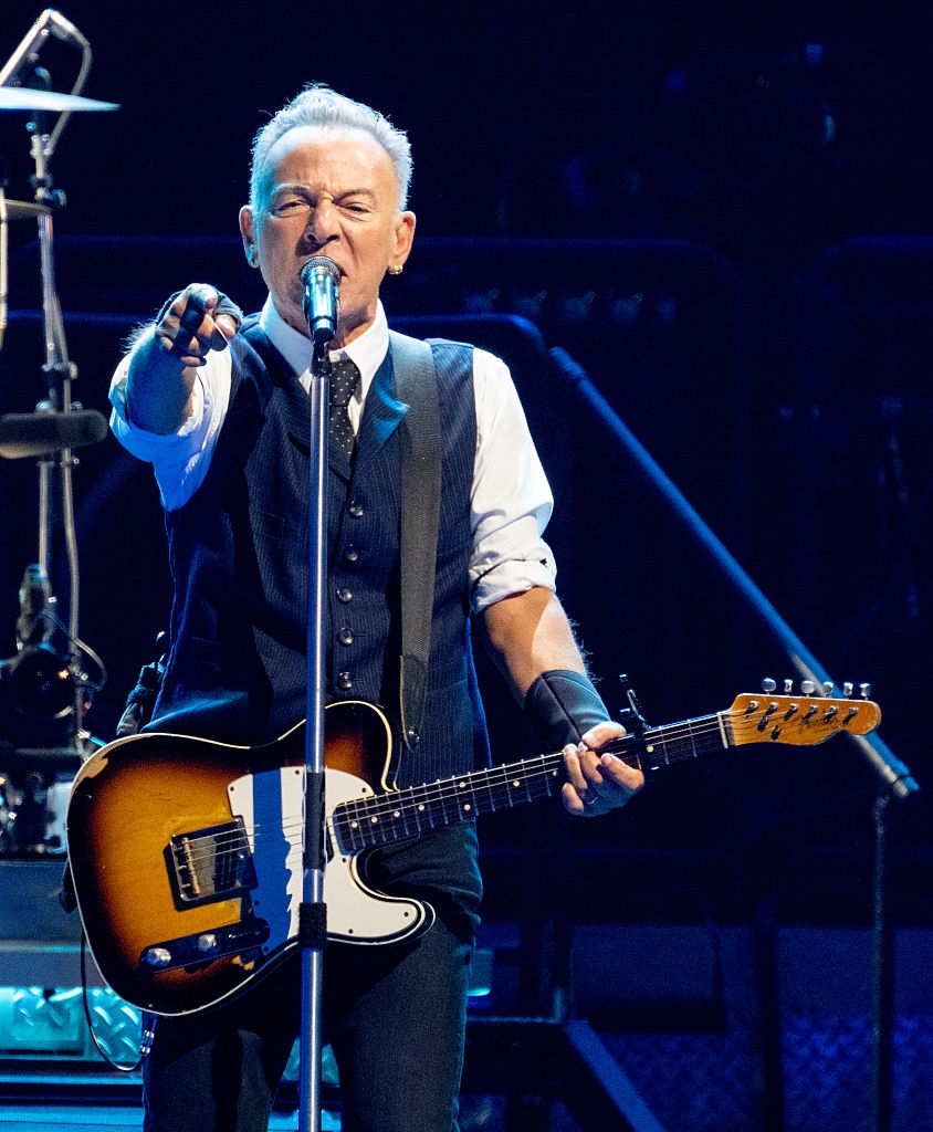 Musician passionately performs onstage, wearing a vest and tie, playing an electric guitar, and engaging with the audience
