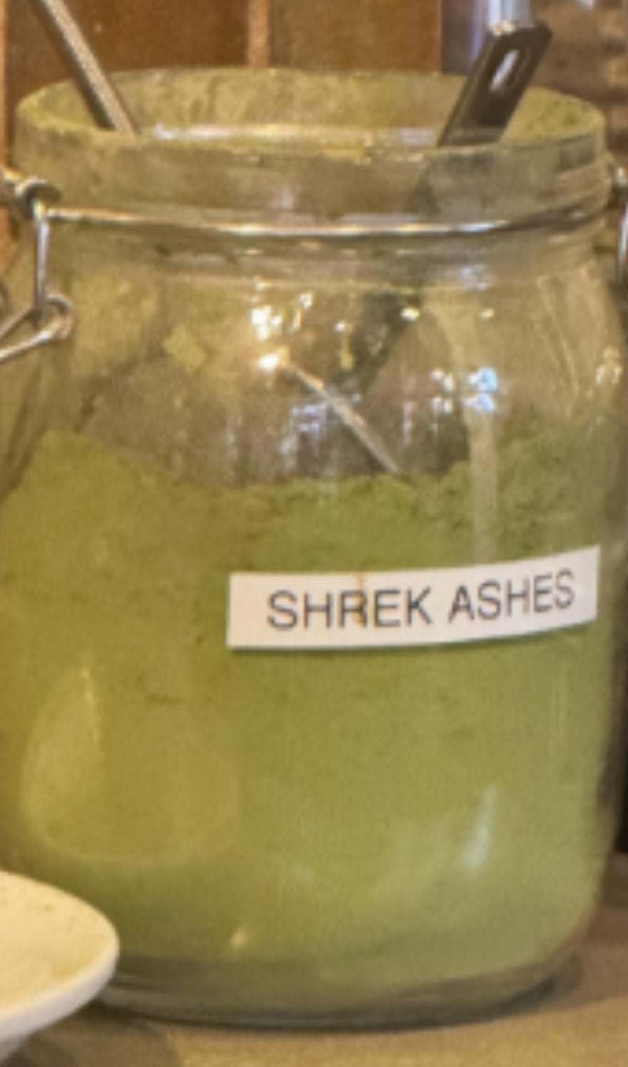 A jar labeled "Shrek Ashes" filled with green powder sits next to a bowl and whisk on a countertop