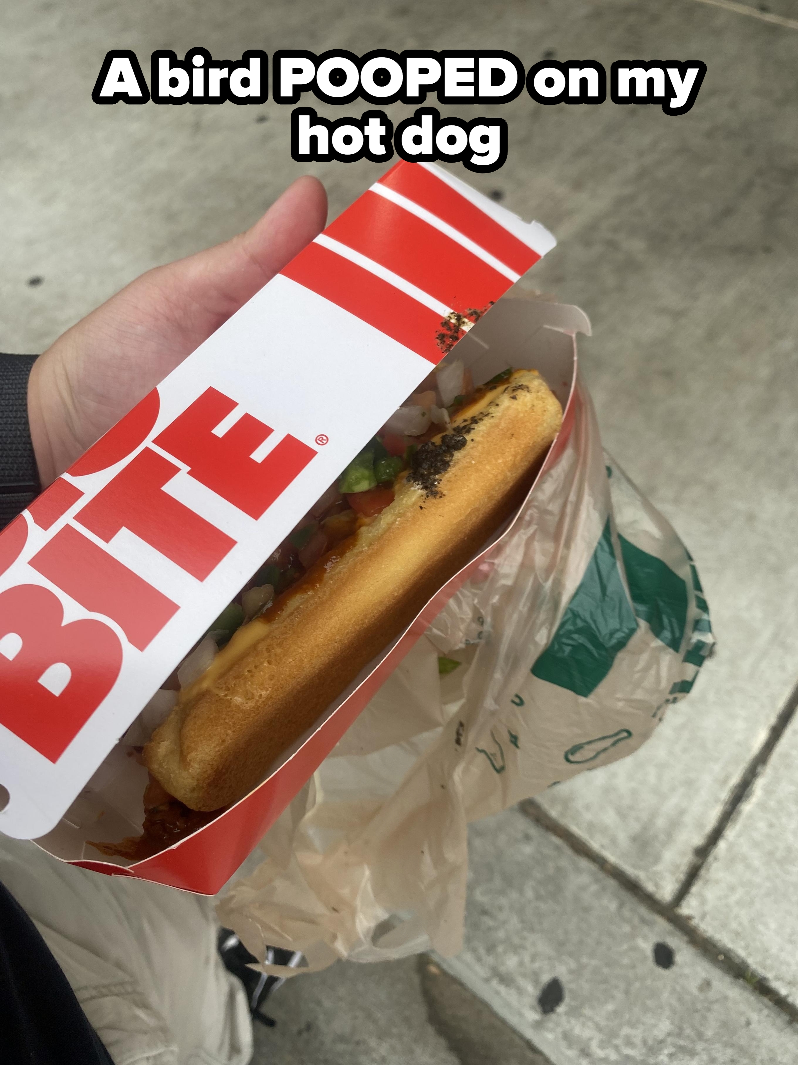 A hand holding a waffle hot dog with toppings, partially in a branded box and a plastic bag on a sidewalk background
