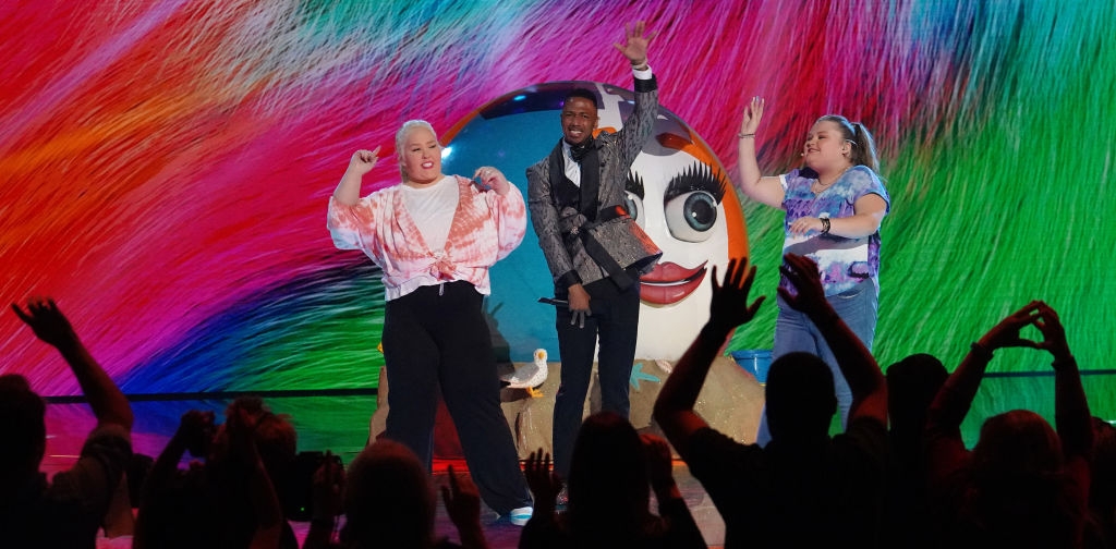 Performers on stage with a colorful backdrop and cheering audience; one wears a patterned jacket, others wear patterned blouses