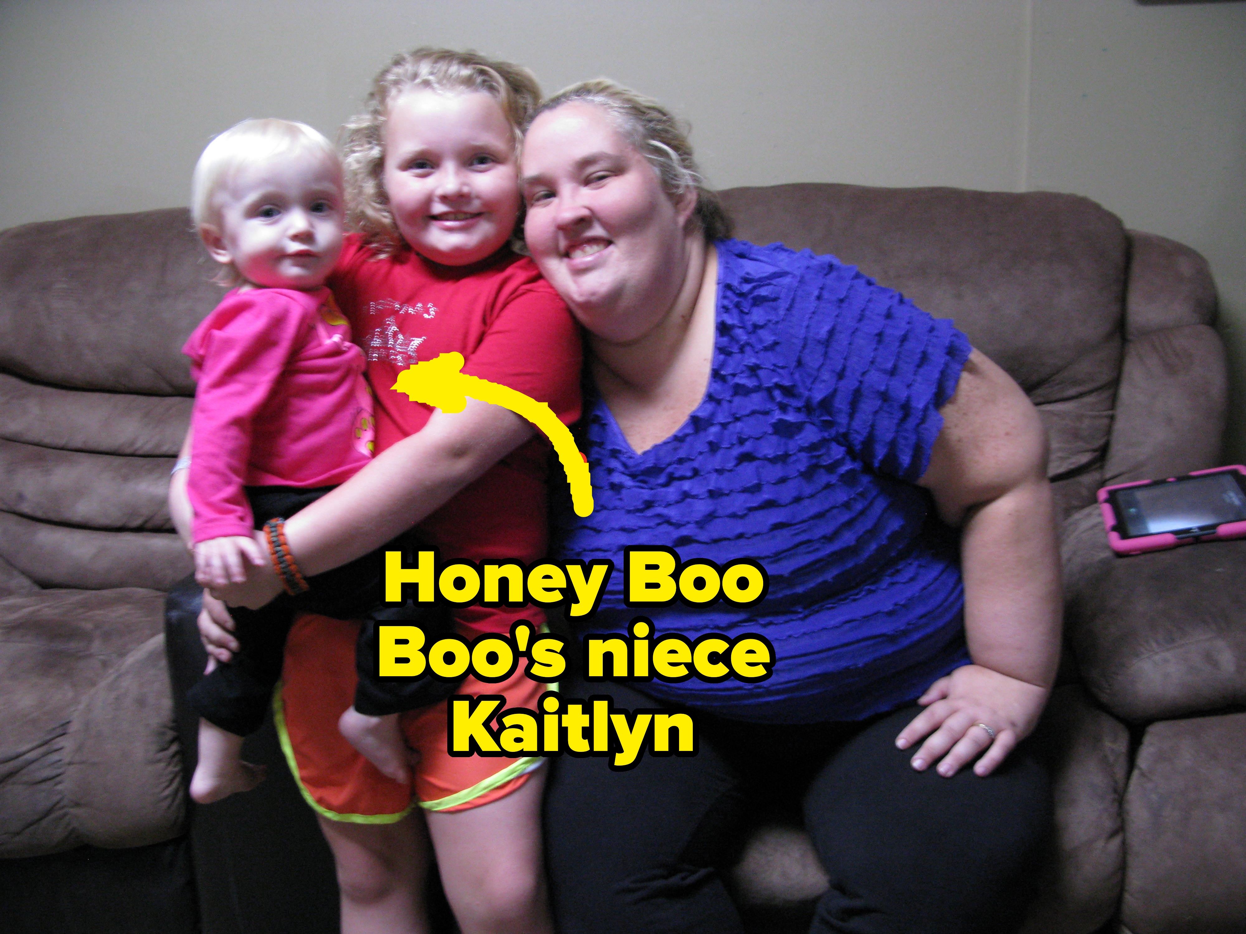 A woman and two children sit on a couch, smiling. One child is standing and the other is sitting on the woman's lap. A tablet rests on the couch