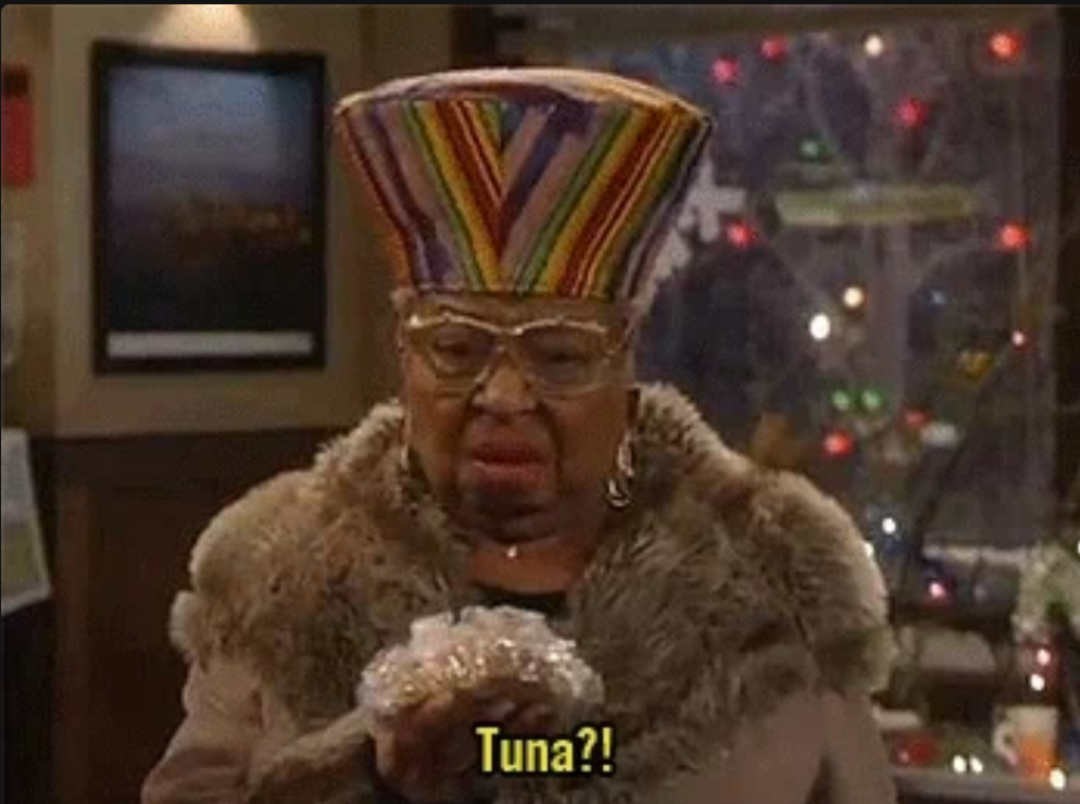 An elderly woman looks at a wrapped sandwich in surprise, exclaiming "Tuna?!" while wearing a unique, tall hat and fur-trimmed coat