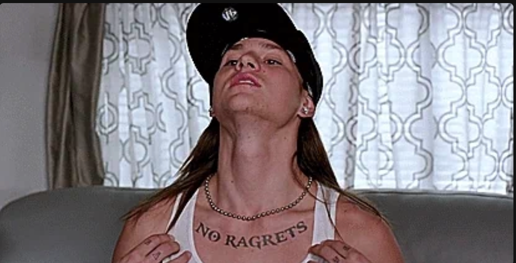 Person showing a "NO RAGRETS" tattoo on chest, wearing a cap backwards and a tank top, looking confidently upward
