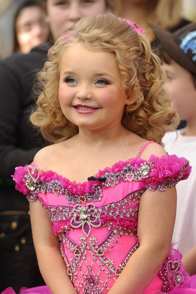 Child in a rhinestone-studded dress smiles at a public event. Curly hair complements the formal style