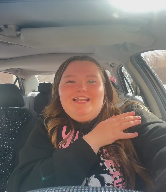 Person in a car, smiling, gesturing with one hand, wearing a casual sweatshirt