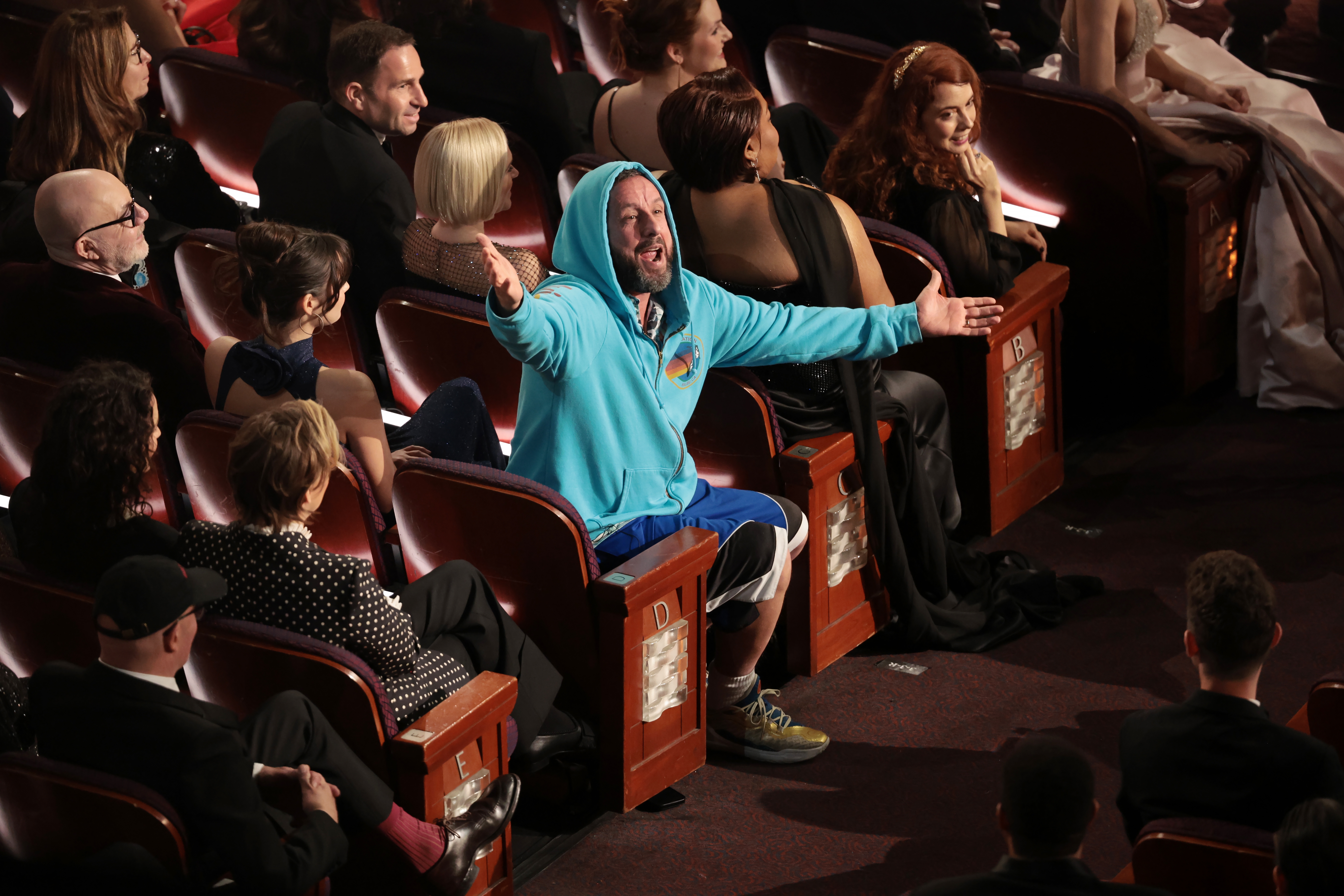 Person in hoodie and shorts stands with arms open among formally dressed audience at an event