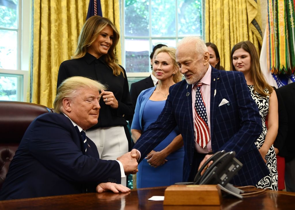 Meeting in the Oval Office; a man in a suit with an American flag tie shakes hands with another man, as others stand nearby, including one woman in a blue dress