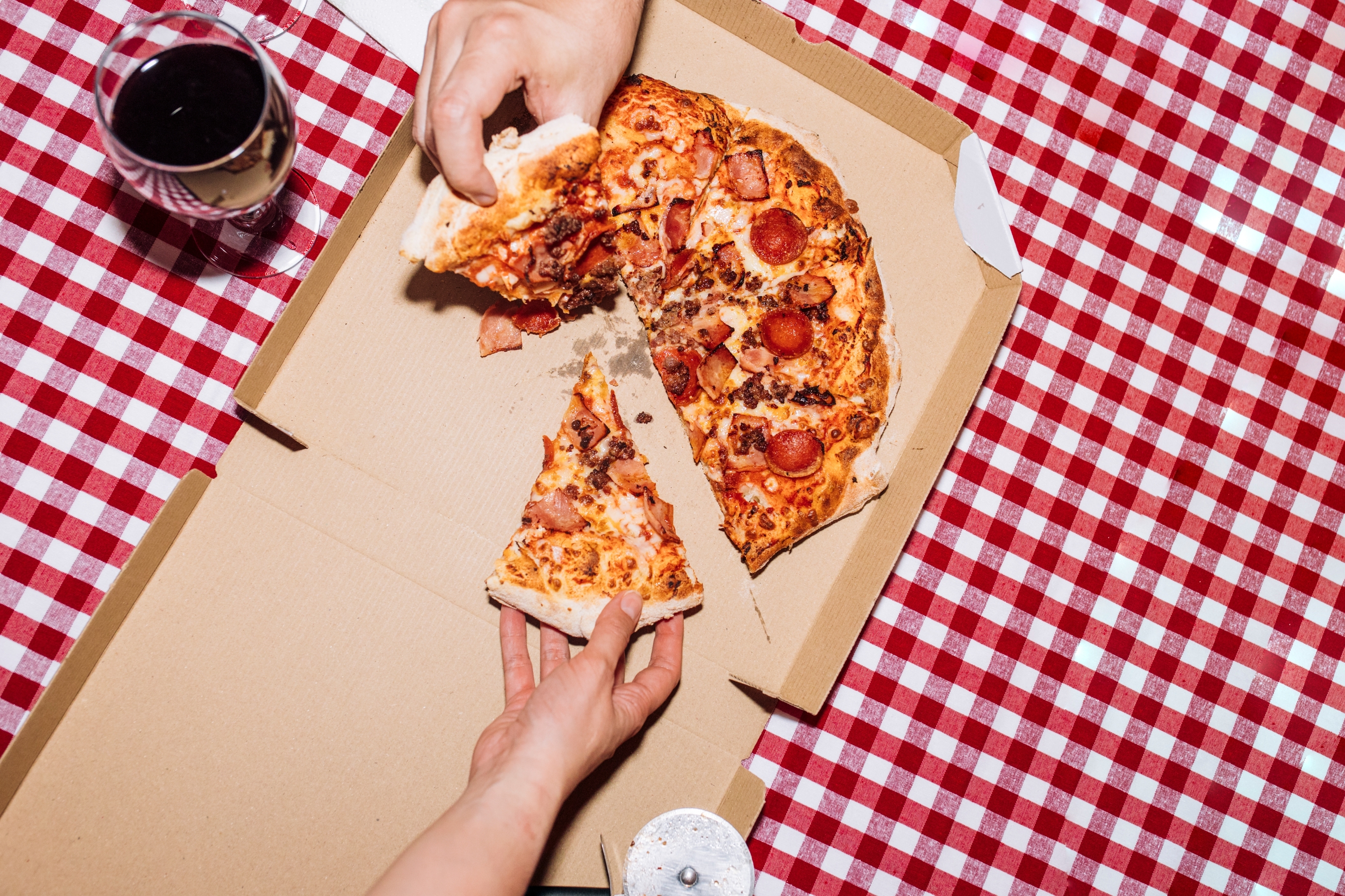Two hands reach for slices from a pizza in a box on a checkered tablecloth, next to a glass of wine