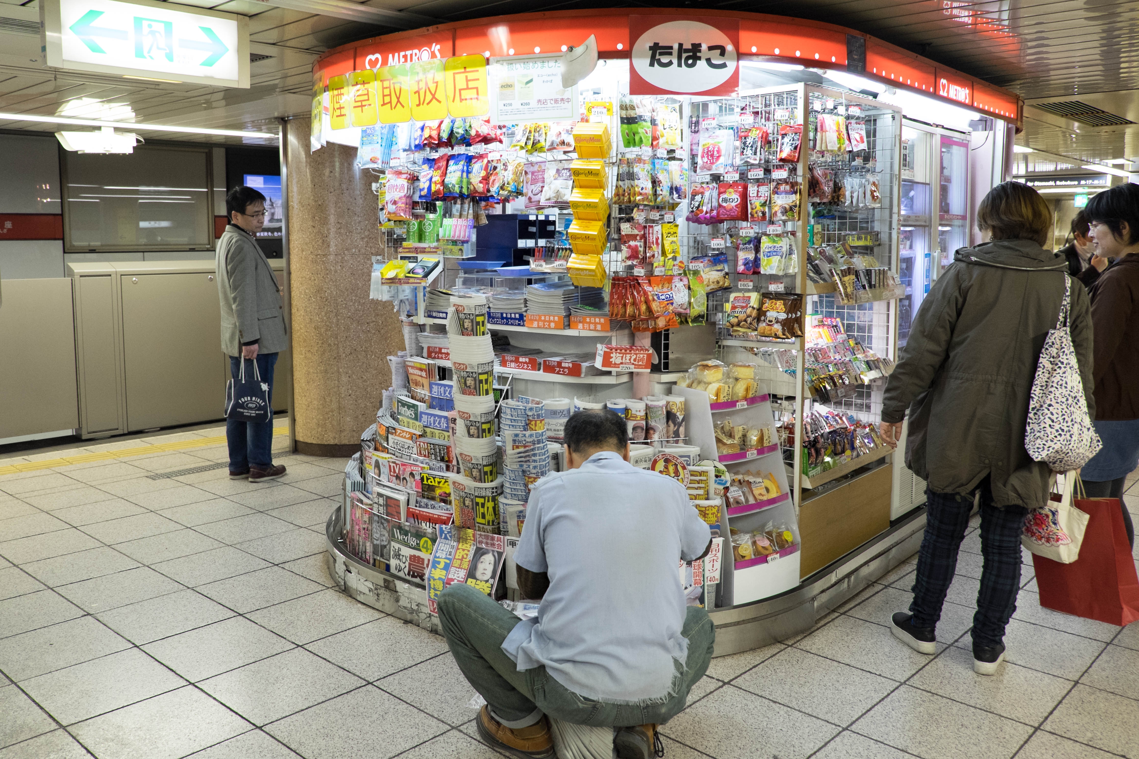 People browsing a newsstand in a train station. Various snacks and magazines are displayed. A person kneels to adjust items on a lower shelf